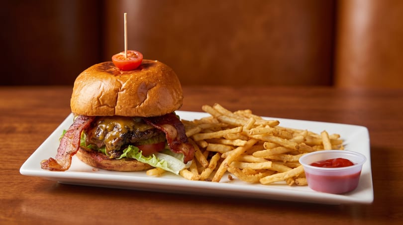 A bacon cheeseburger with lettuce, tomato, and a side of french fries with ketchup on a white plate.